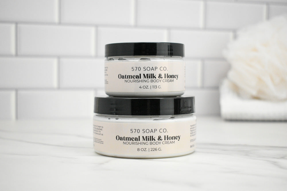 Two jars of Oatmeal Milk & Honey body cream from 570 Soap Co. on a white surface with a tiled wall background.