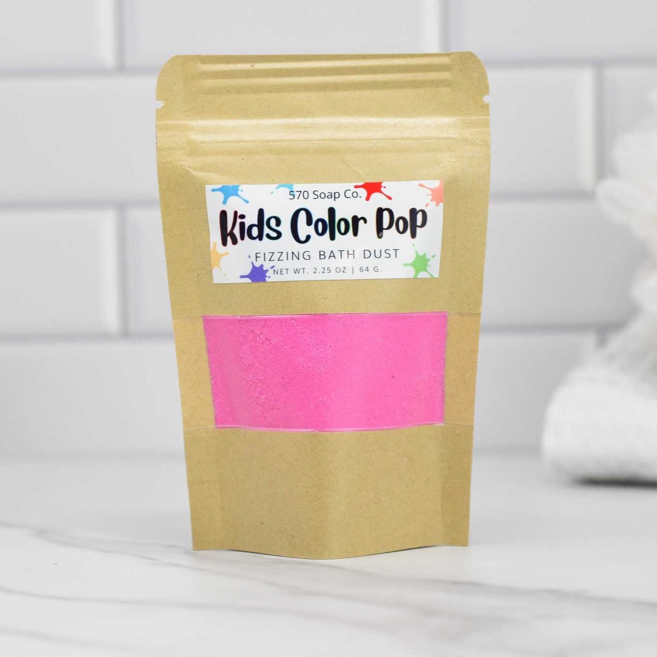 A standing pouch of Kids Color Pop Fizzing Bath Dust by 570 Soap Co. promises a vibrant adventure with its pink bath dust, highlighted against a white tiled background with a plush white towel on the right.