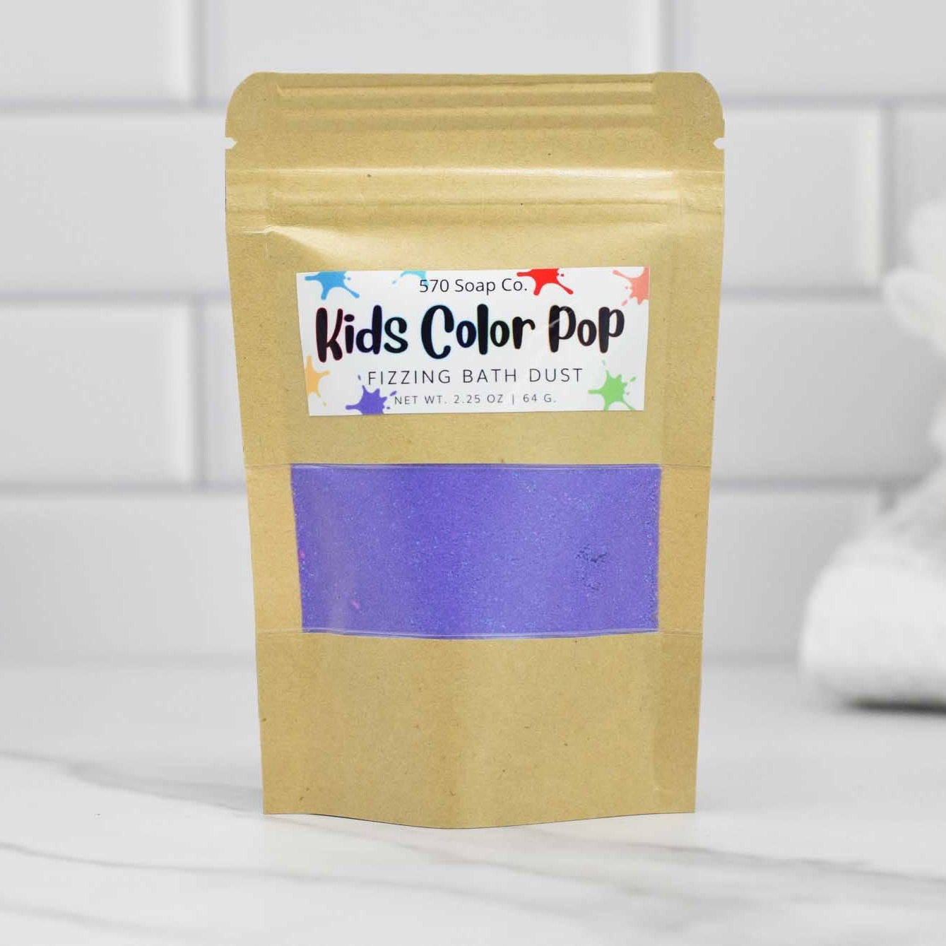 A brown paper bag labeled 570 Soap Co.'s Kids Color Pop Fizzing Bath Dust rests on a white marble surface, revealing the vibrant blue powder inside through its translucent window. Against the tiled wall backdrop, it promises a Vibrant Adventure.