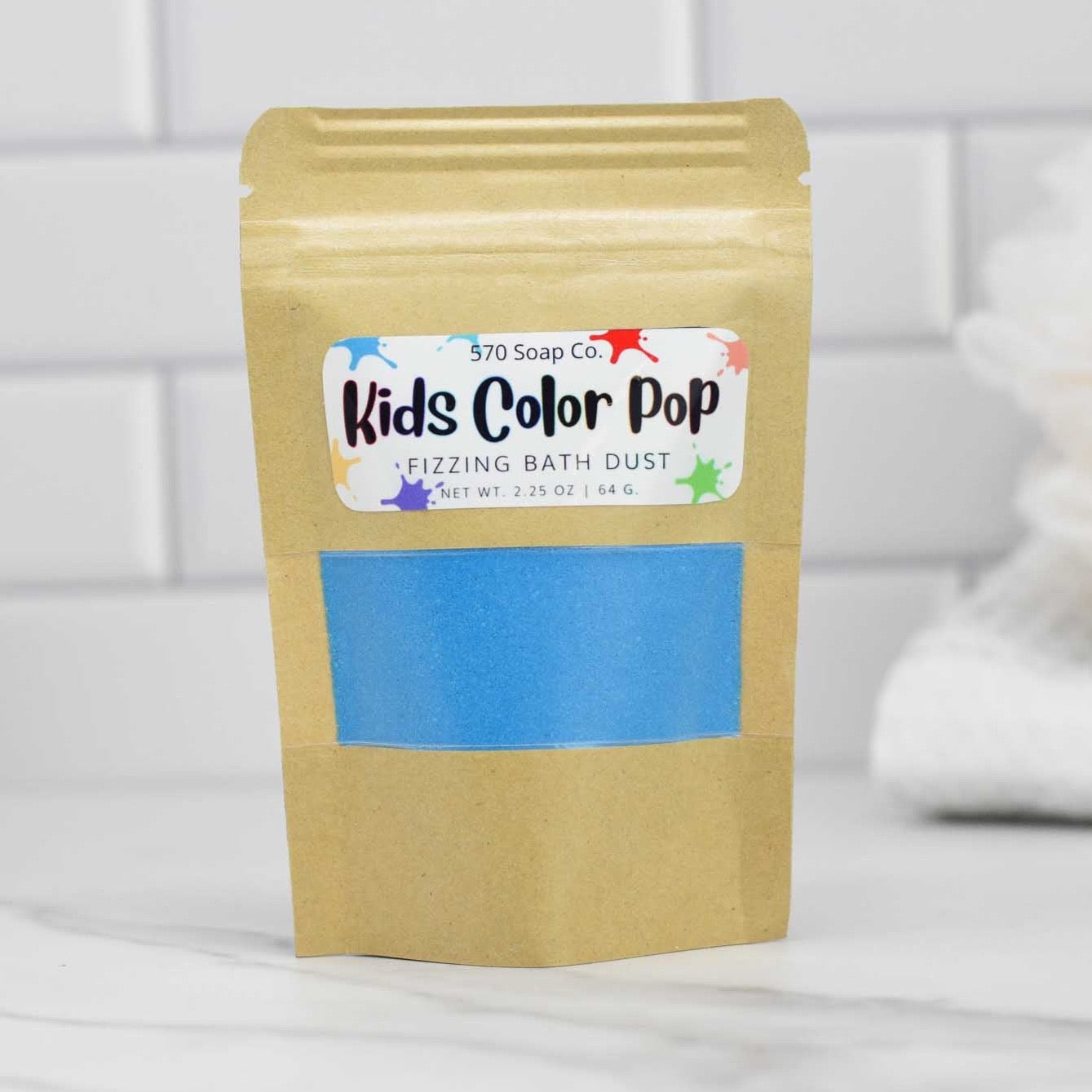 A brown paper bag branded by 570 Soap Co., showcasing the Kids Color Pop Fizzing Bath Dust with a clear window, reveals a vibrant adventure in blue powder inside. Situated on a white marble surface against a backdrop of white tiles, it promises an exciting, colorful bath experience for kids.