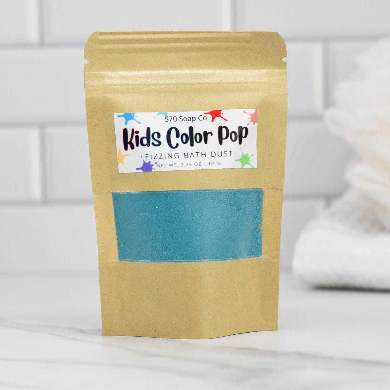 A tan pouch of 570 Soap Co.'s Kids Color Pop Fizzing Bath Dust is displayed on a bathroom counter, promising a vibrant adventure. The package reveals blue bath dust through a clear window, complemented by fresh white towels and a sleek tiled wall in the background.