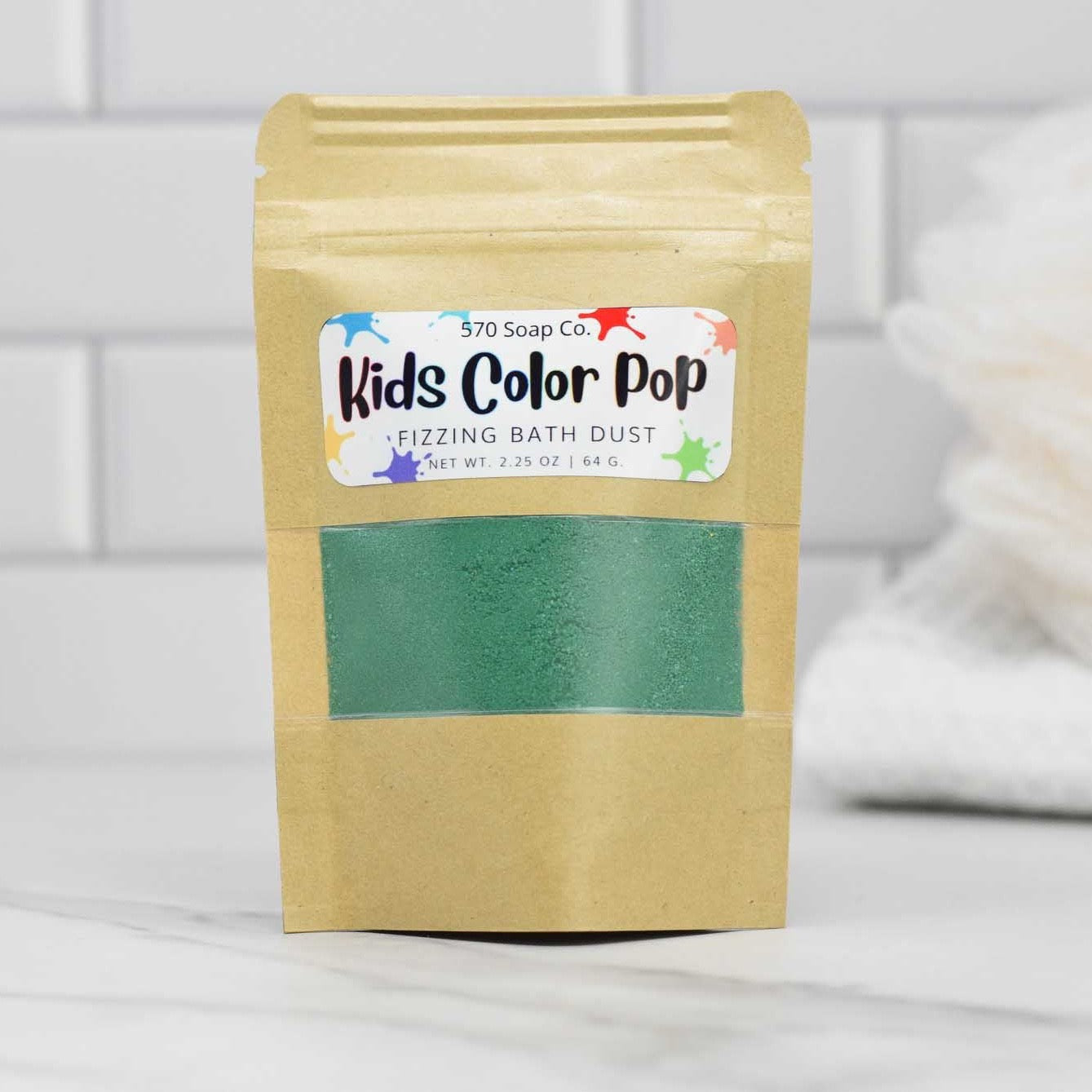 A brown paper package from 570 Soap Co. labeled Kids Color Pop Fizzing Bath Dust showcases a see-through section brimming with vibrant green powder. Placed on a white tiled surface, it invites little ones to an exciting adventure, with towels neatly stacked in the background.
