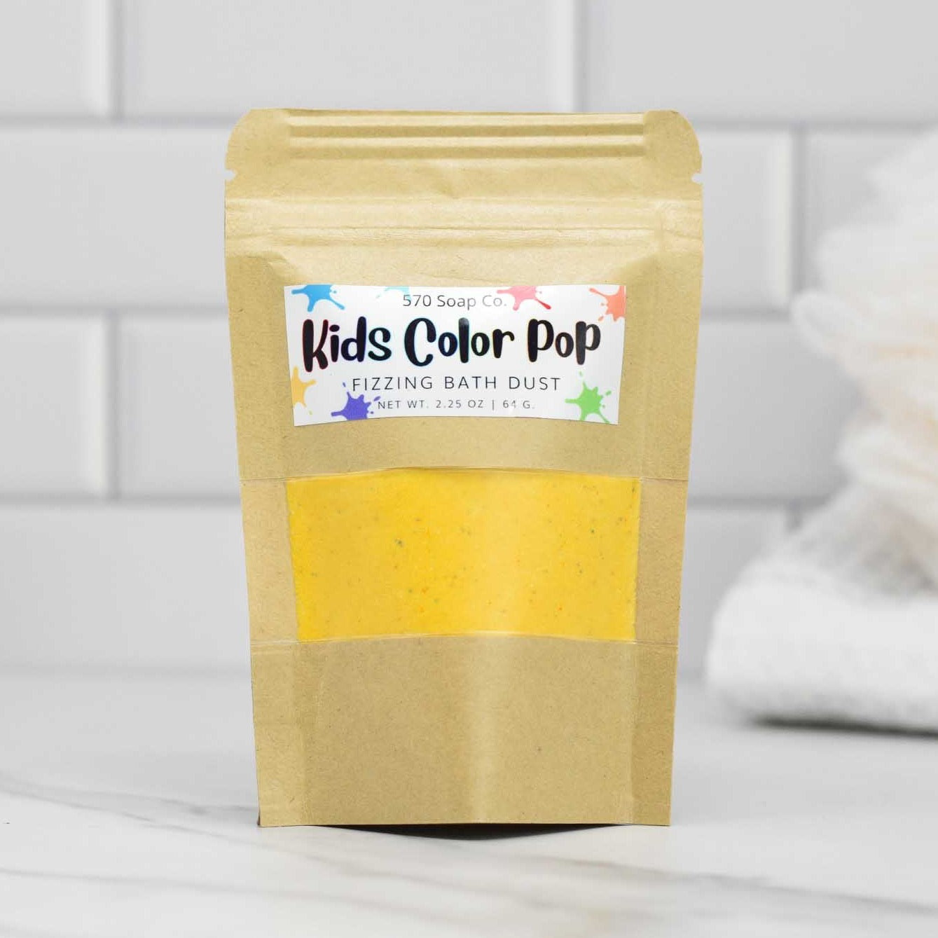 A package of 570 Soap Co.'s Kids Color Pop Fizzing Bath Dust sits on a countertop, offering a vibrant bath-time adventure. The brown packaging features a colorful label and a clear window that reveals the bath dust inside, with a white towel enhancing the scene in the background.