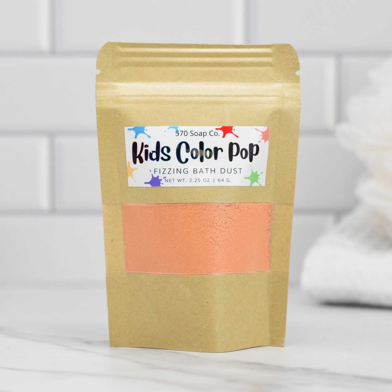 A 2.25-ounce (64g) package of Kids Color Pop Fizzing Bath Dust from 570 Soap Co. sits on the bathroom counter, hinting at a lively and colorful experience ahead. The packaging features a brown hue adorned with a vibrant label, while a soft white towel rests out of focus in the background, anticipating cozy post-bath cuddles.