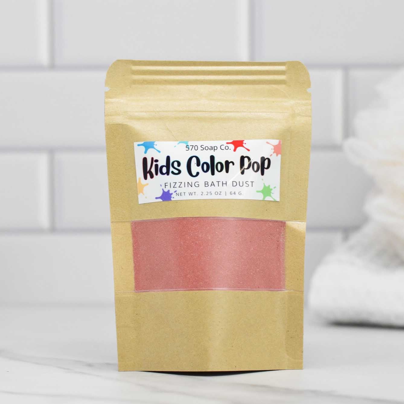 A standing brown paper package, labeled Kids Color Pop Fizzing Bath Dust from 570 Soap Co., holds red bath dust. The bag sits in front of a white tile backdrop, accompanied by a pristine white towel.