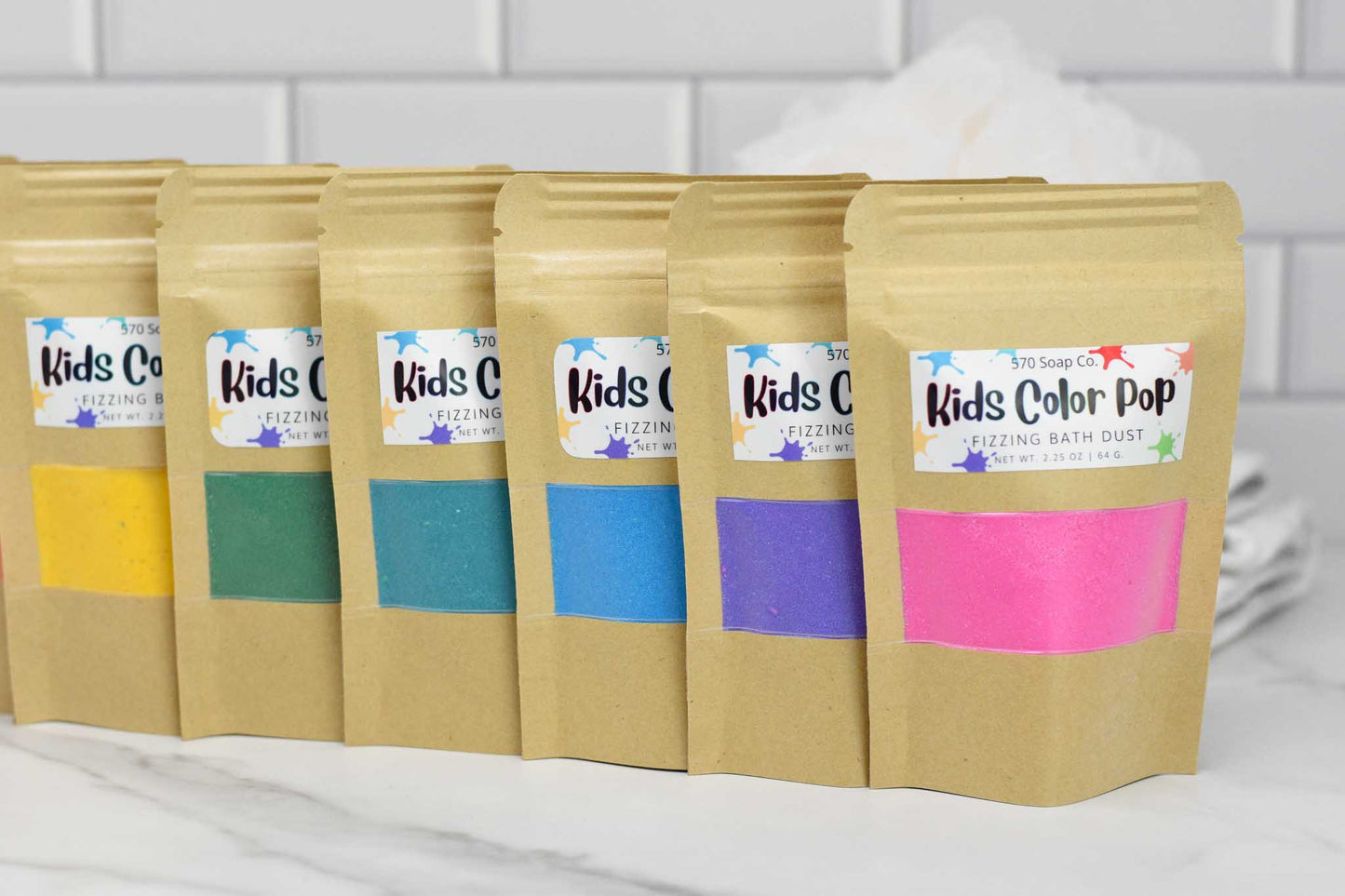 A row of six brown paper bags from 570 Soap Co., featuring Kids Color Pop Fizzing Bath Dust, promises a vibrant adventure with fizzing bath dust in vivid shades of yellow, green, teal, blue, purple, and pink. These bags stand out against a crisp white tiled background.