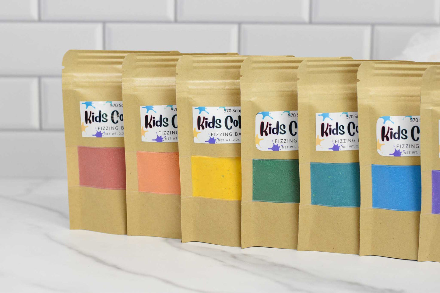 Seven brown paper bags with colorful labels are displayed against a white tiled background. Each label features the name "570 Soap Co." and showcases a different colored band: pink, orange, yellow, green, blue, teal, and purple. These vibrant Kids Color Pop Fizzing Bath Dust bags promise an exciting adventure inside.