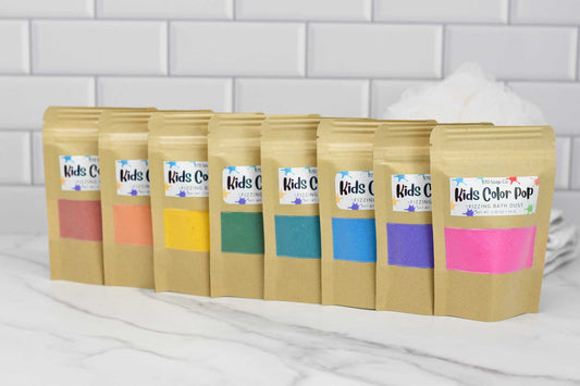 A row of eight small brown paper bags from 570 Soap Co., each labeled with "Kids Color Pop Fizzing Bath Dust," promises a vibrant adventure with its colorful fizzing bath dust. The bags, featuring hues such as red, orange, yellow, green, blue, teal, purple, and pink, are neatly lined up on a marble surface.