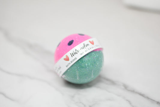 A round bath bomb from 570 Soap Co. rests on a marble surface, featuring a pink top with black dots that mimic a watermelon, and a green bottom. A label around the middle reads "Watermelon Bath Bomb: Round," weighing 5.4 oz / 170 g, evoking summer nostalgia with its skin-loving formula.