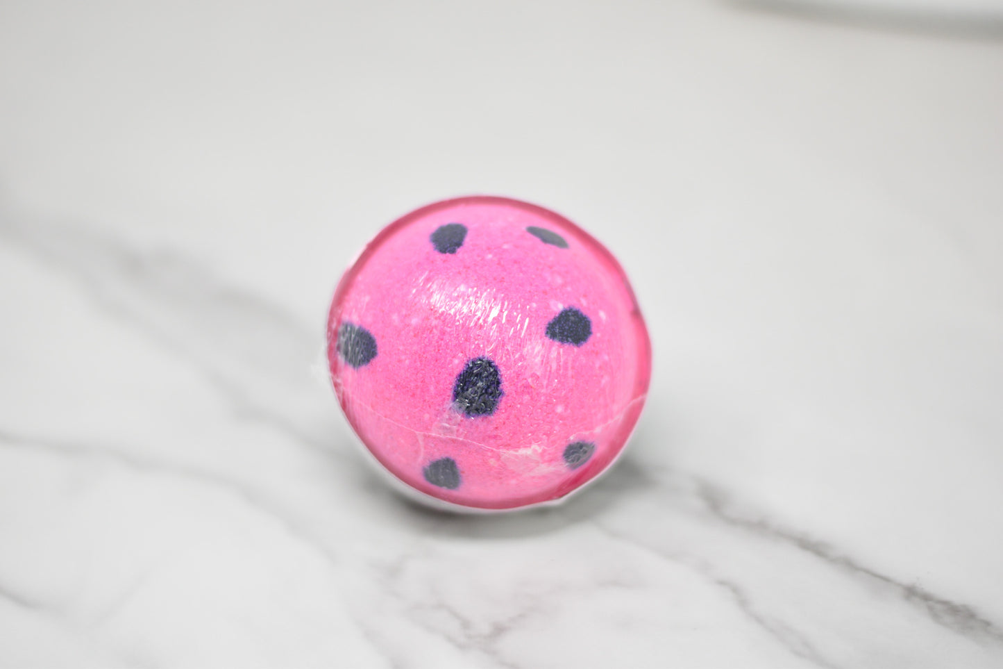 A round Watermelon Bath Bomb from 570 Soap Co., in a delightful pink with black polka dots, rests on a smooth, white marbled surface. Its glossy finish evokes summer nostalgia while hinting at its skin-loving formula.