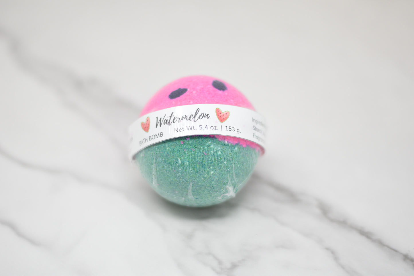 The Watermelon Bath Bomb: Round from 570 Soap Co. is a two-tone delight with a pink top and green bottom, capturing the essence of summer nostalgia. A white label encircles its middle, adorned with text and small heart designs. Positioned on a marble surface, this bath bomb delivers a skin-loving formula for the ultimate pampering experience.
