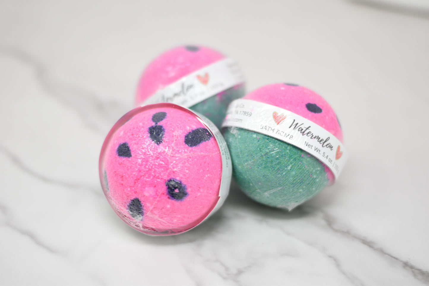 Three Watermelon Bath Bombs from 570 Soap Co., featuring a pink and green design with black speckles, evoke summer nostalgia on a white marble surface. Each round bath bomb is wrapped in clear plastic labeled with "Watermelon" and contains a skin-loving formula for the ultimate refreshing soak.