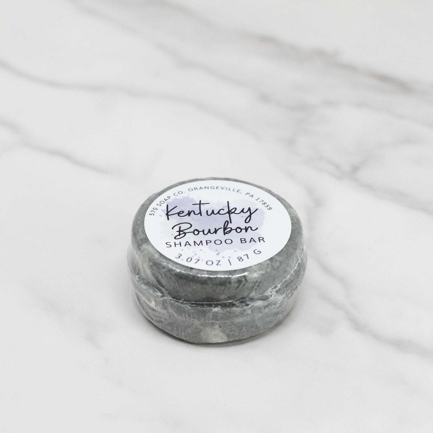 A round soap or shampoo bar from 570 Soap Co., labeled as the Kentucky Bourbon Shampoo Bar, sits on a marble surface. The gray bar is adorned with a white circular label displaying black and blue text. Infused with bourbon fragrance oil, it provides a luxurious lather. The packaging states the product weight as 3.07 oz.