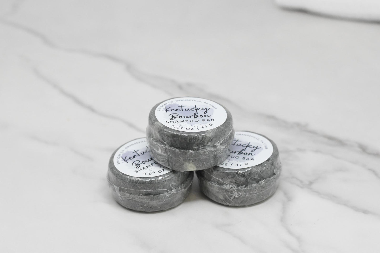 Three elegantly stacked gray shampoo bars, infused with a touch of bourbon fragrance oil, rest on a light marble surface. Each 3-ounce Kentucky Bourbon Shampoo Bar by 570 Soap Co. offers a luxurious lather for an indulgent bathing experience.