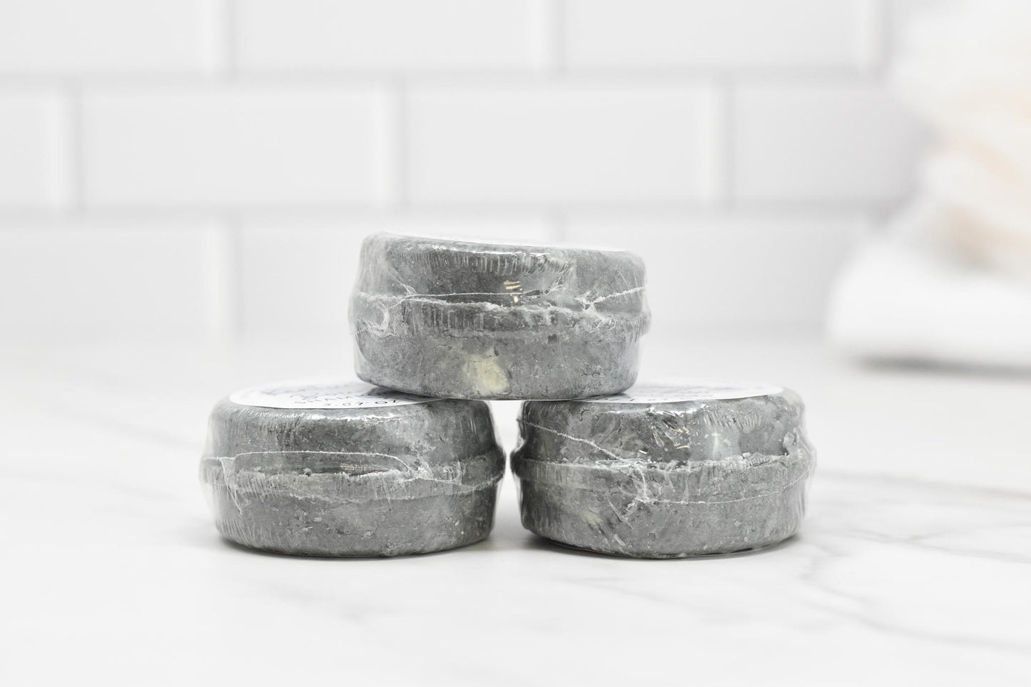 Three Kentucky Bourbon Shampoo Bars by 570 Soap Co., wrapped in clear plastic, are stacked on a white marble surface. They exude a subtle note of bourbon fragrance oil. The background features white subway tiles, enhancing the minimalist and clean aesthetic of the scene while hinting at a luxurious lather experience.