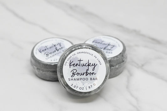 Three gray Kentucky Bourbon Shampoo bars are elegantly displayed on a white marbled surface, offering a luxurious lather and infused with bourbon fragrance oil. Each bar from 570 Soap Co. in Orangeville, PA, weighs 3.07 ounces (87 grams) and features a sleek white and black design.