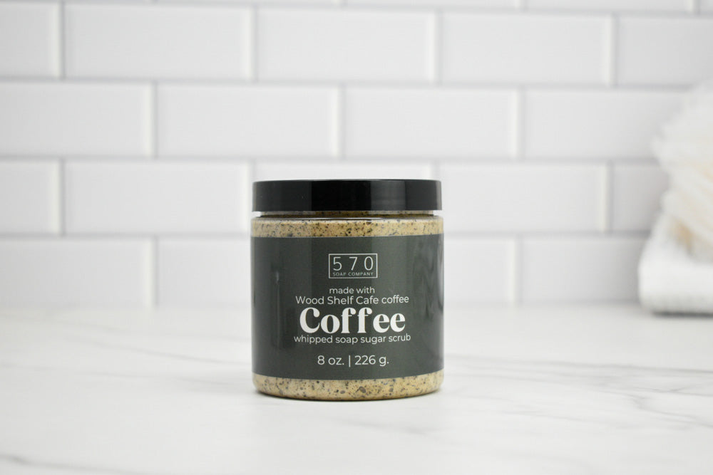 Jar of coffee-flavored whipped soap sugar scrub on a white surface with a tiled wall background