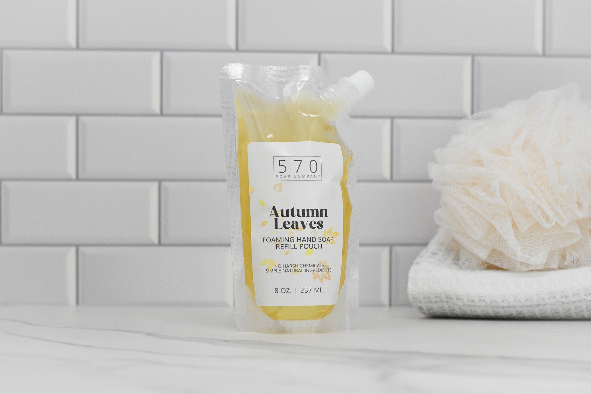 An 8 oz (237 mL) refill pouch of Autumn Leaves Scented Foaming Hand Soap from 570 Soap Co., with its delightful Fall Season Aroma, is placed on a countertop against a white tile backsplash. Nearby, you can see a white shower sponge and folded washcloths.
