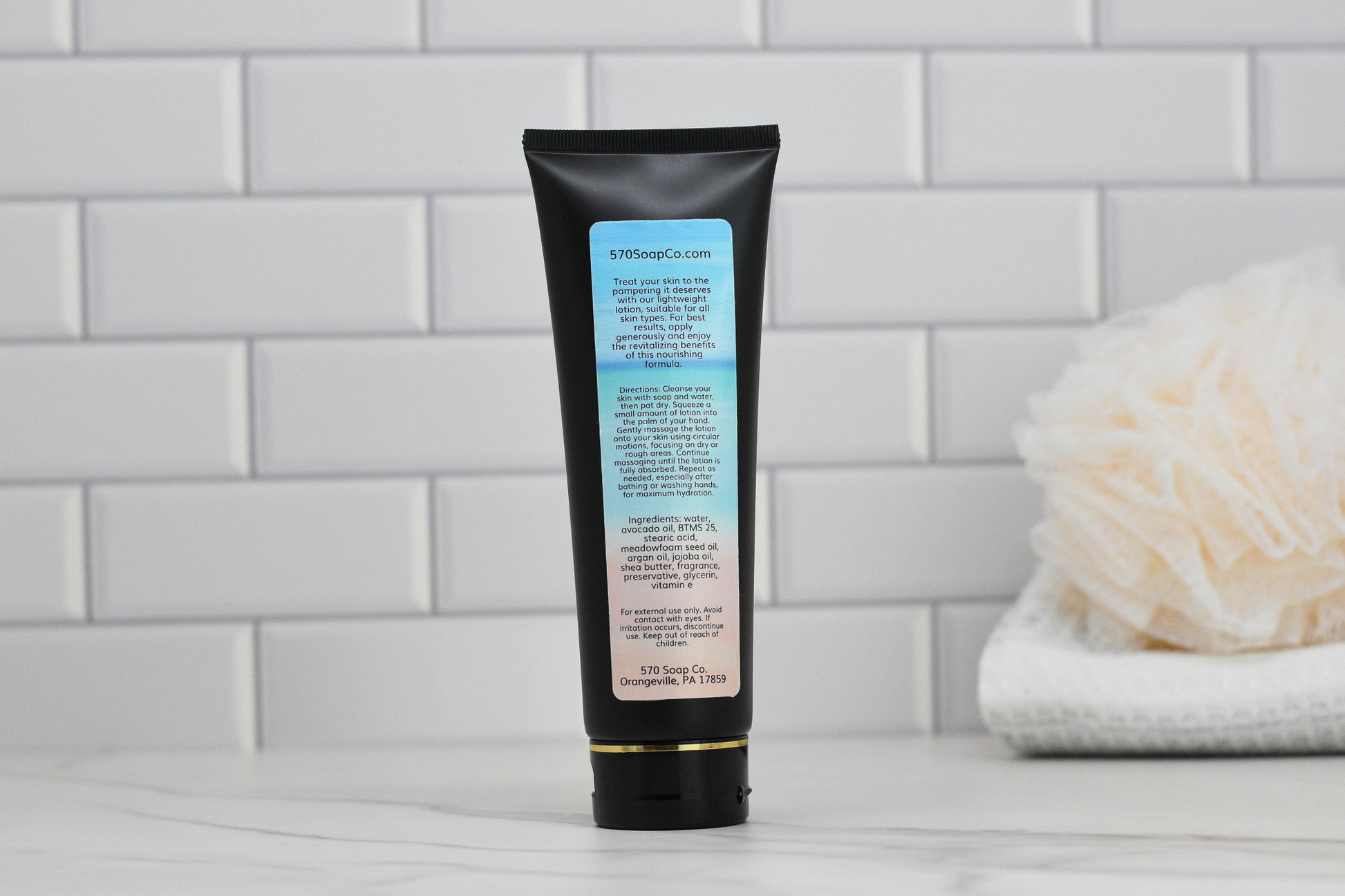 A black tube of Beach Vibes Lightweight Lotion by 570 Soap Co. stands upright on a marble countertop against a backdrop of white subway tiles, with the back label text in clear view. To add texture to this exquisite skincare scene, a white loofah is placed nearby, hinting at the nourishing qualities of its skin-loving ingredients.