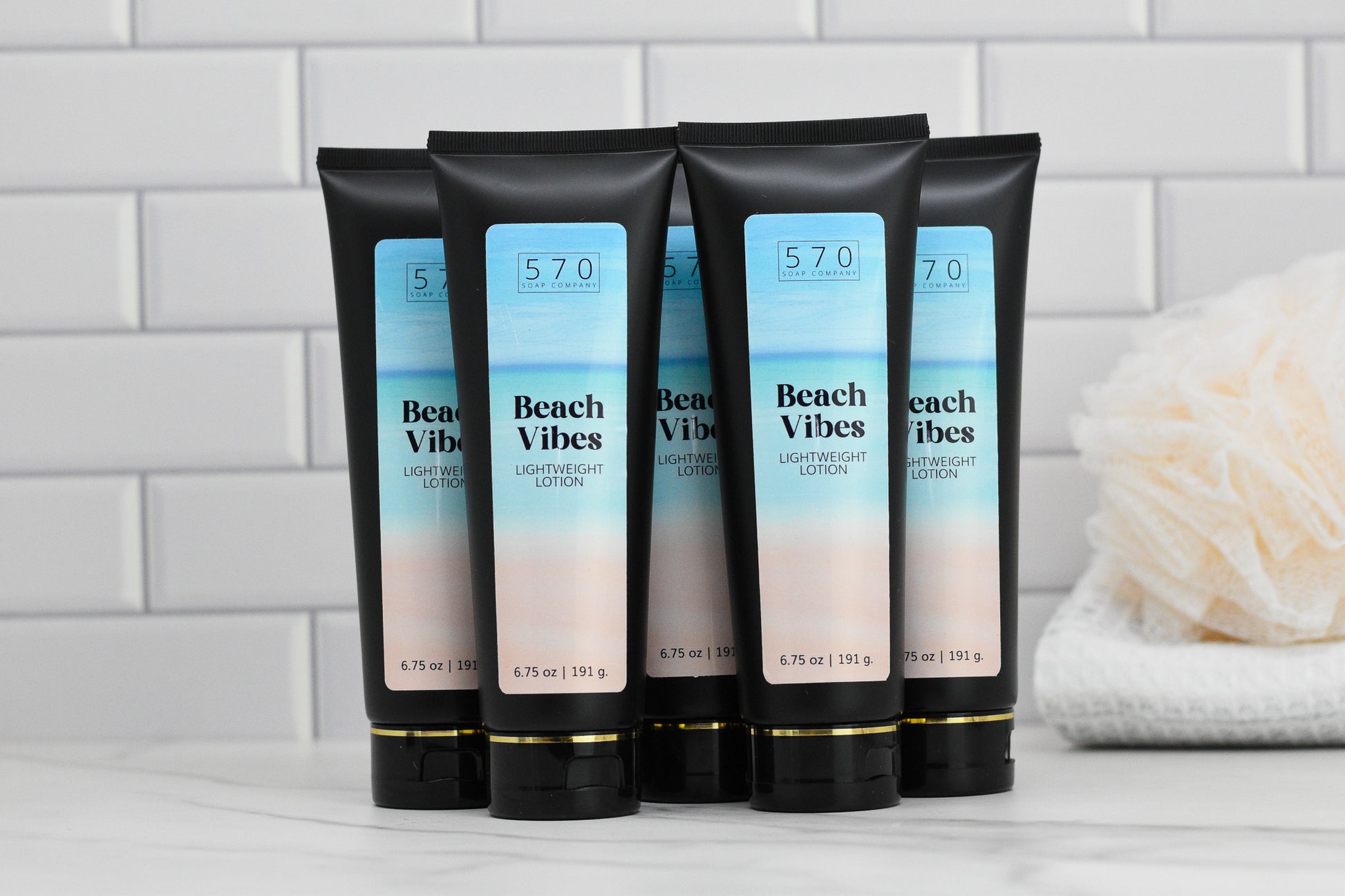 Six tubes of Beach Vibes Lightweight Lotion by 570 Soap Co. are neatly arranged in two rows on a white surface. Infused with skin-loving ingredients, the packaging boasts a stunning gradient design transitioning from blue to beige. In the background, a white bath sponge rests against a white tiled wall.
