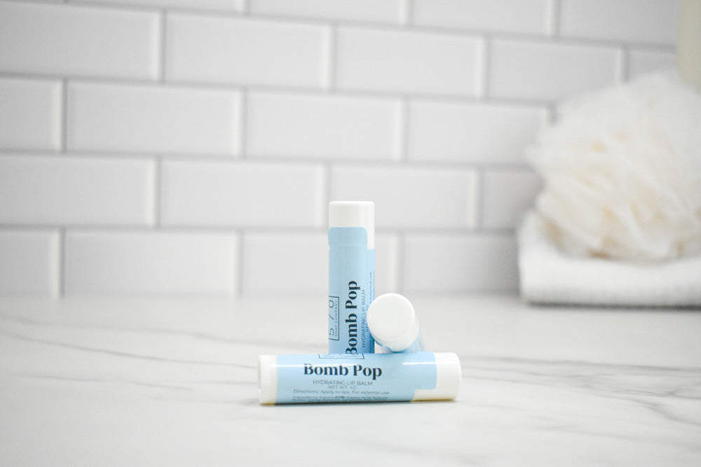 Two lip balm tubes labeled 'bomb Pop' on a bathroom counter with a tiled wall background.