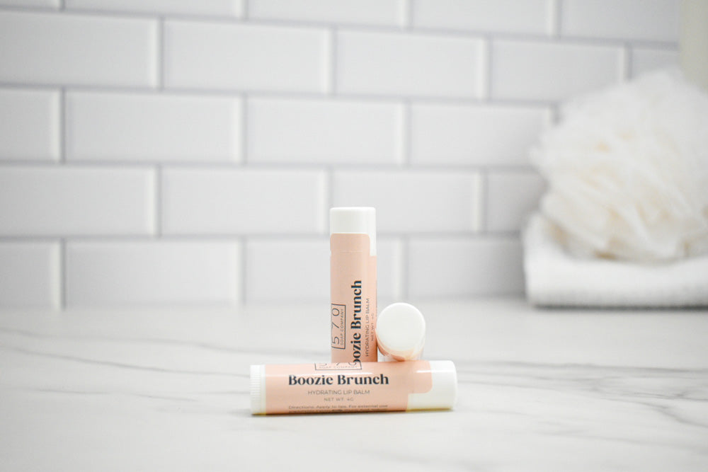 Two lip balms with 'Boozie Brunch' branding on a white surface with a tiled wall background.