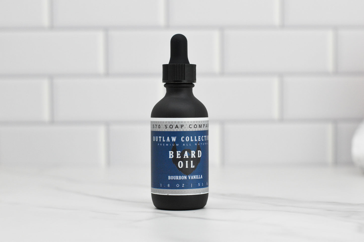 A bottle from the Beard Oil - Choose your scent collection by 570 Soap Co. is placed on a white tiled surface. This dark bottle is enriched with natural oils for optimal beard care and comes with a dropper cap and a striking blue label, featuring the scent Bourbon Vanilla from their Outlaw Collection under Premium Male Grooming.