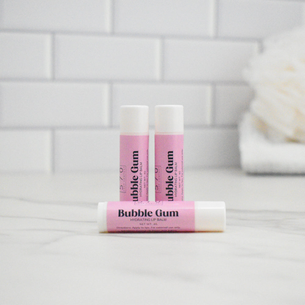Three lip balms labeled 'Bubble Gum' on a white surface with a tiled wall background.