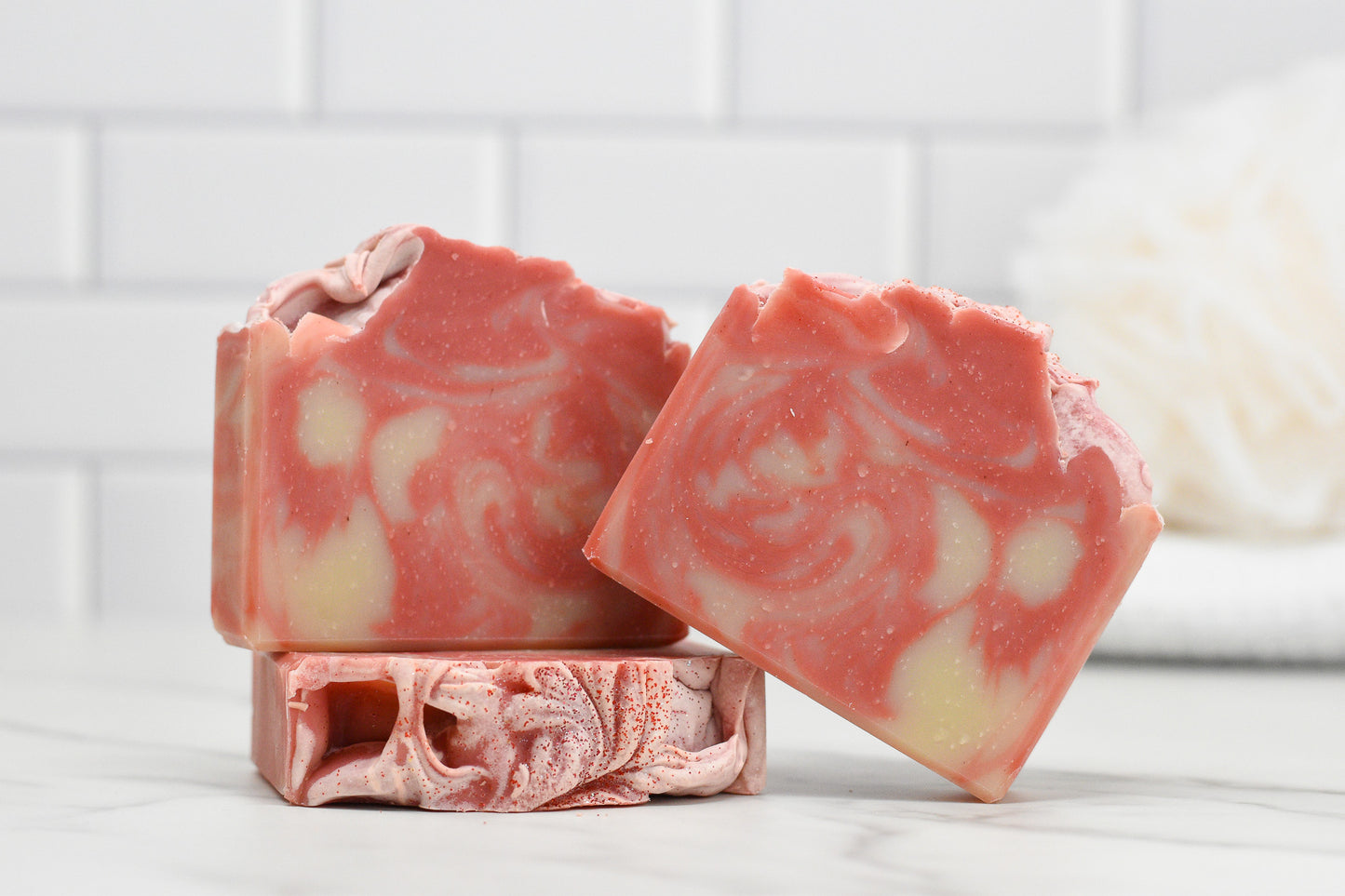Three bars of 570 Soap Co.'s Candy Cane Lane Bar Soap are displayed against a white tiled background. One stands upright, while the other leans, highlighting its marbled design and twisted peppermint essence. A blurred bath sponge in the background hints at the soap's moisturizing formula.