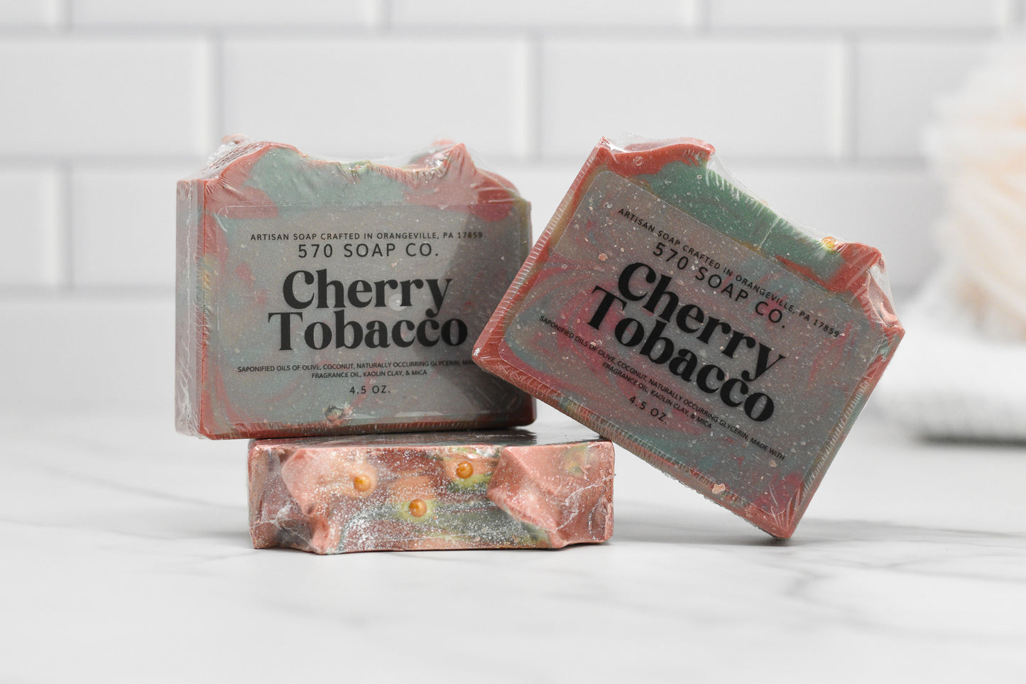 Three bars of Cherry Tobacco Bar Soap from 570 Soap Co. are shown, infused with calming kaolin clay. The soap showcases a marbled pattern with swirls of green, red, and gold on a white tiled surface, accompanied by a blurred shower puff in the background.