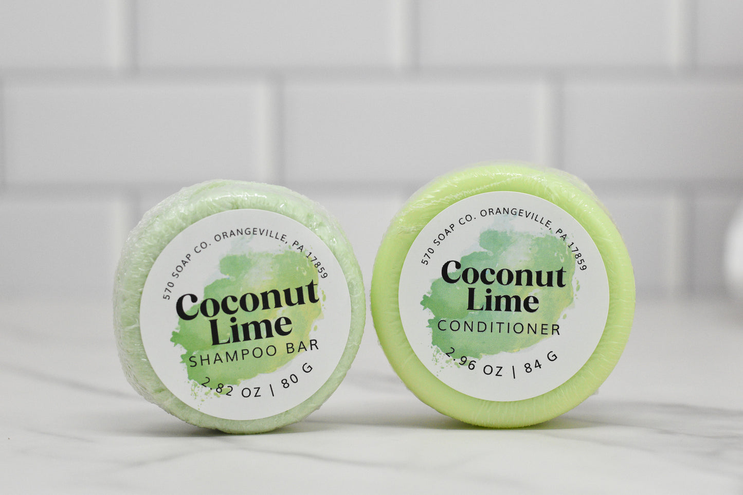 Positioned side by side on a white surface are two round bars from 570 Soap Co.: the Coconut Lime Shampoo Bar, weighing 2.82 oz, and the Conditioner Bar for All Hair Types - Coconut Lime, weighing 2.96 oz. They rest against a backdrop of subway tiles.