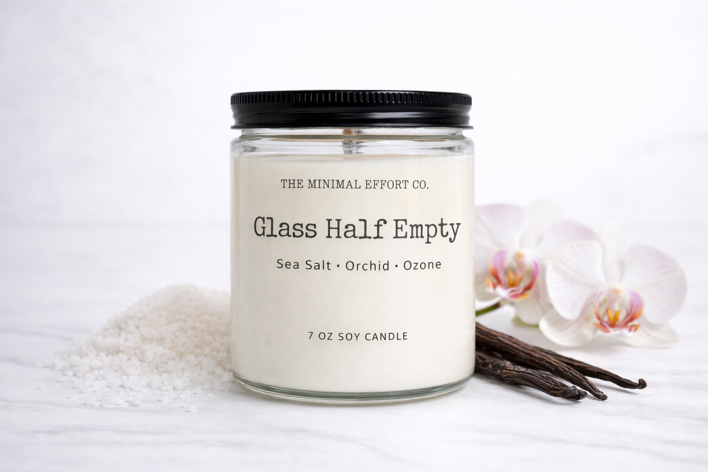 Candle labeled 'Glass Half Empty' from 'The Minimal Effort Co.' with vanilla beans and orchids on a white background