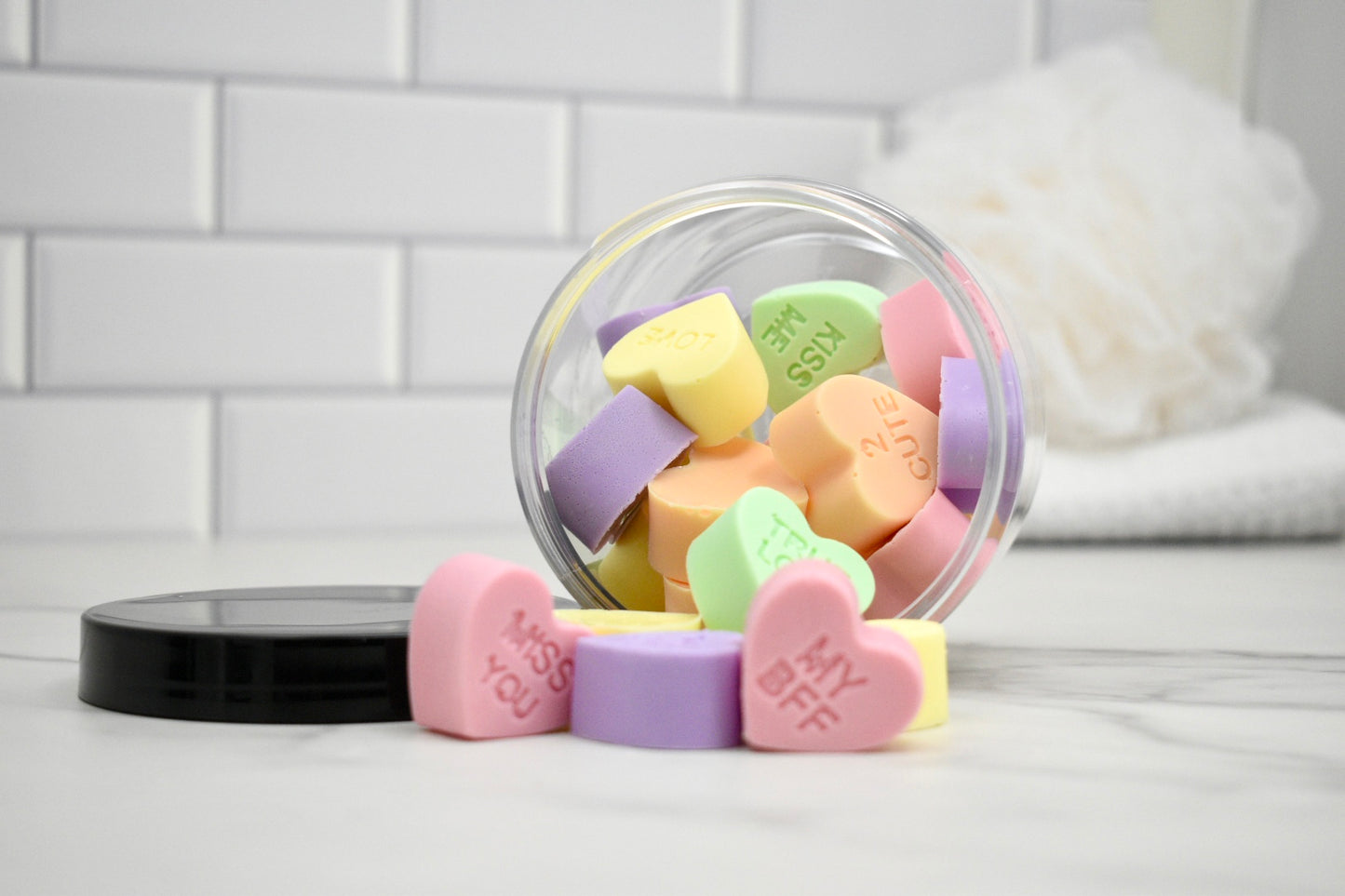 Colorful heart-shaped candy pieces spilling out of a clear jar on a white surface with a blurred background.