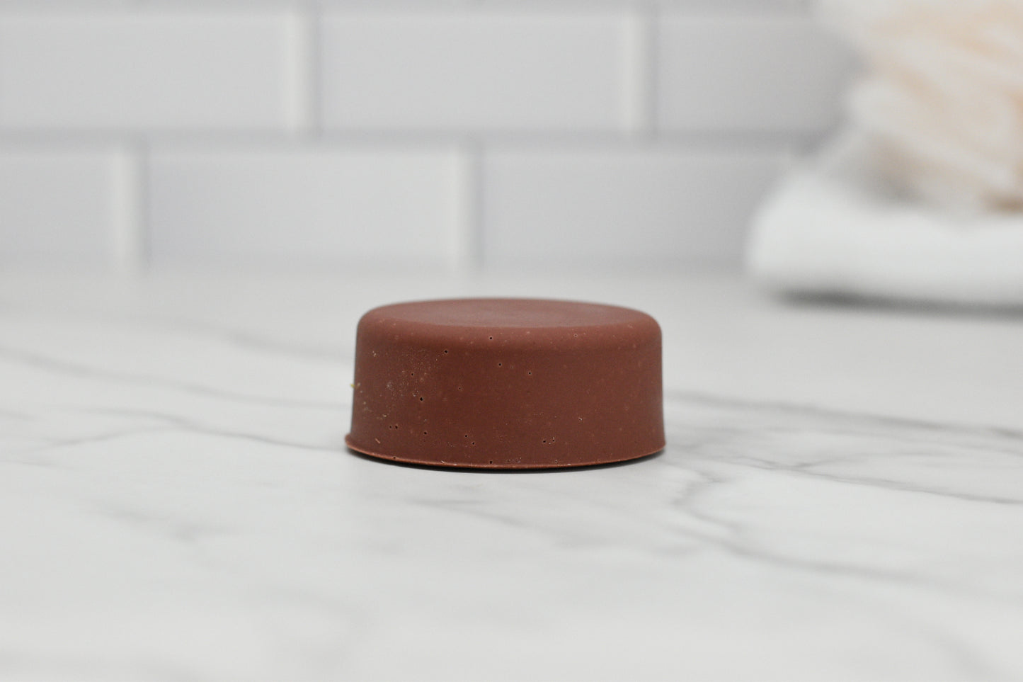 A compact, round bar of solid brown Dog Shampoo Bar - All Natural by 570 Soap Co. with a soothing lavender scent is placed on a marble countertop. The background highlights white subway tile and a blurred beige towel.