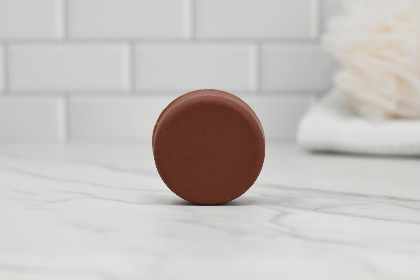 A circular brown bar of 570 Soap Co.'s Dog Shampoo Bar - All Natural rests upright on a white marble surface, with a blurred white towel in the background and a white tiled wall behind it.