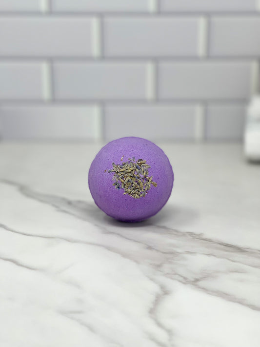 A Lavender Bath Bomb by 570 Soap Co., infused with essential oil, rests on a marble countertop, offering a calming touch of aromatherapy against the white tiled backsplash.