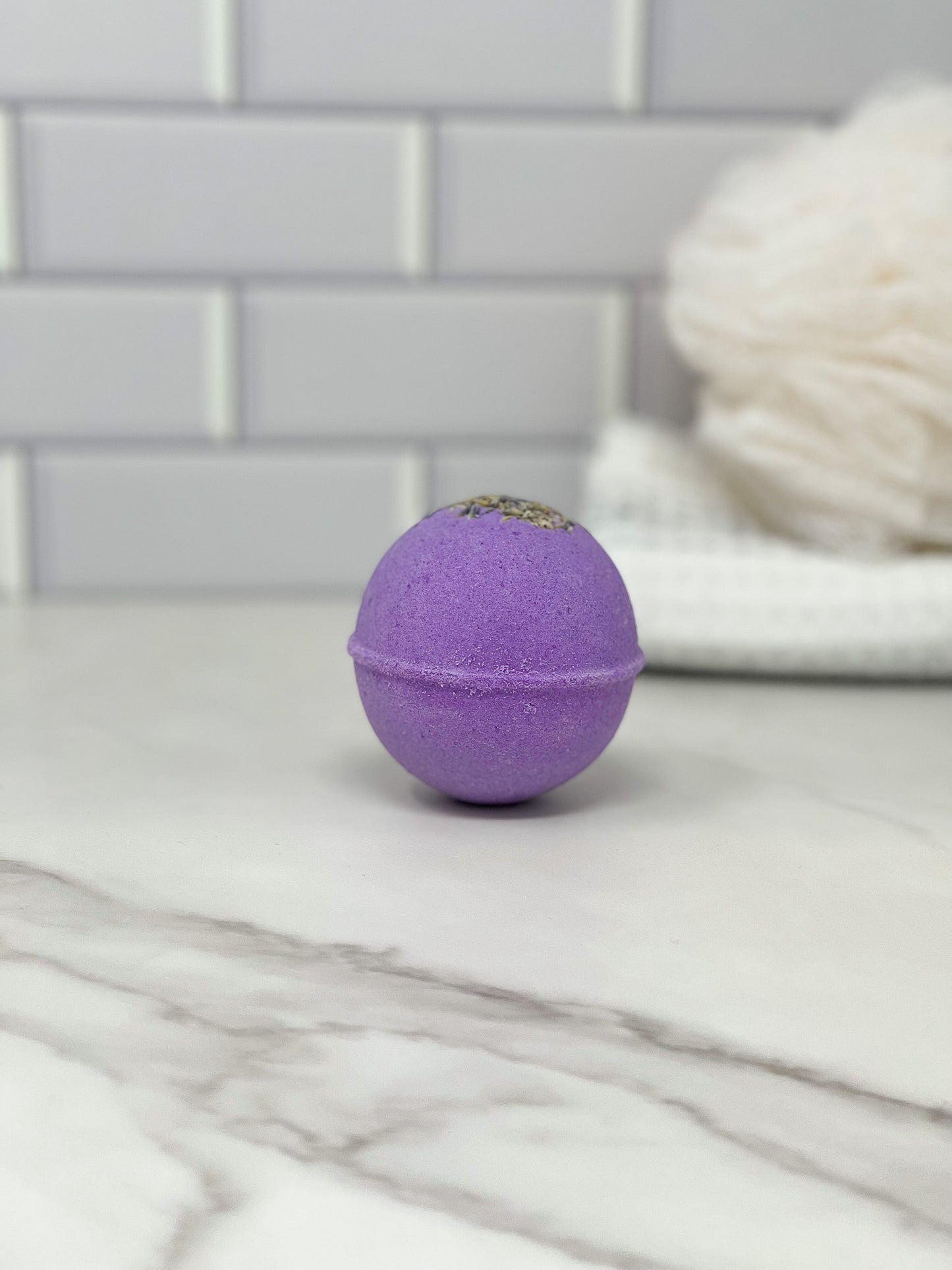The **Lavender Bath Bomb - Made with Essential Oil** by **570 Soap Co.** rests on a marble countertop against a white, textured background. Aromatherapy elements seamlessly merge into the setting, accompanied by a folded white towel that adds a calming touch to the softly blurred background.