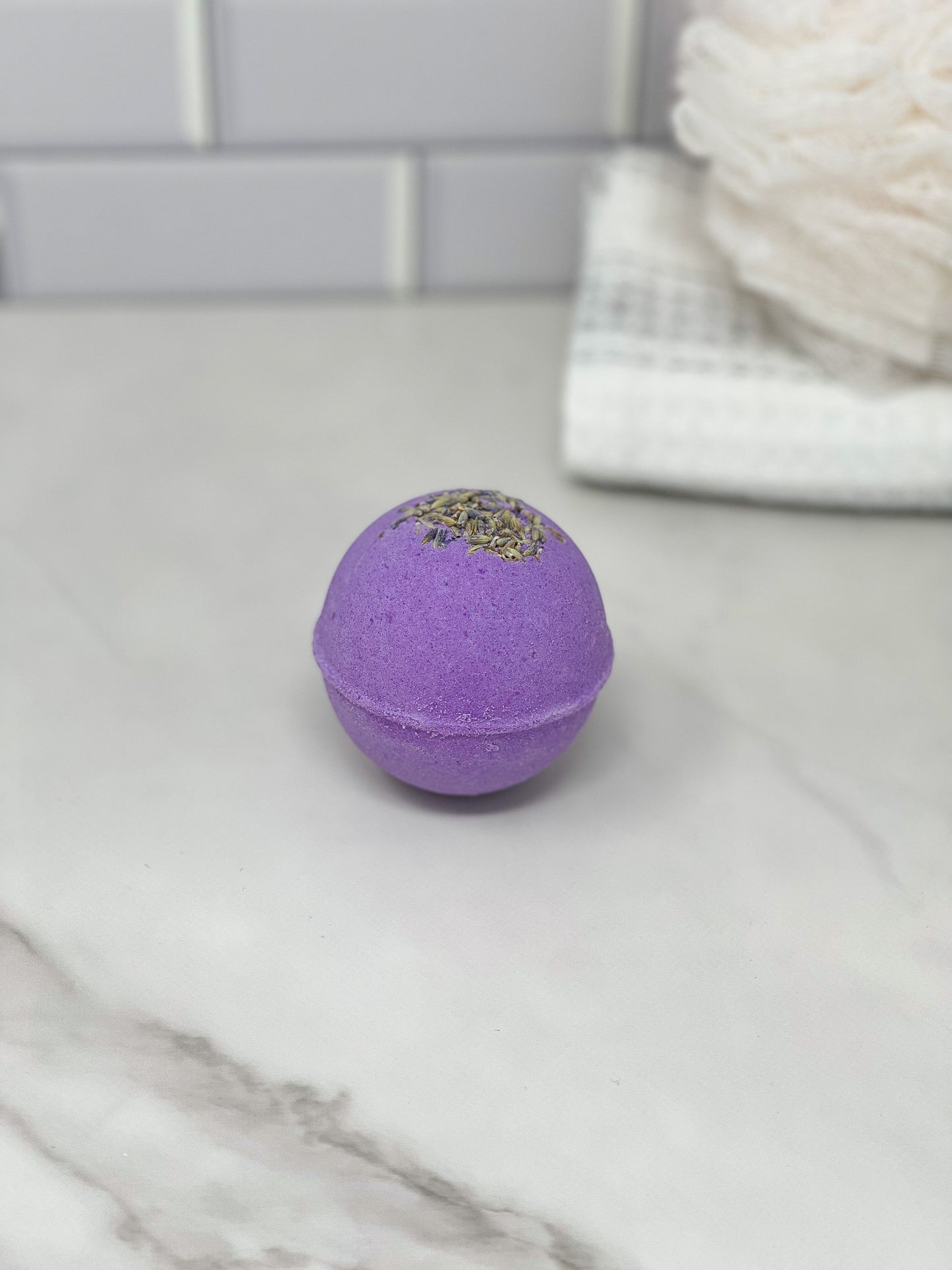 A Lavender Bath Bomb by 570 Soap Co., crafted with essential oil and adorned with dried lavender, rests on a marble surface. In the background, a white tiled wall and a stack of towels create an aromatherapy oasis.