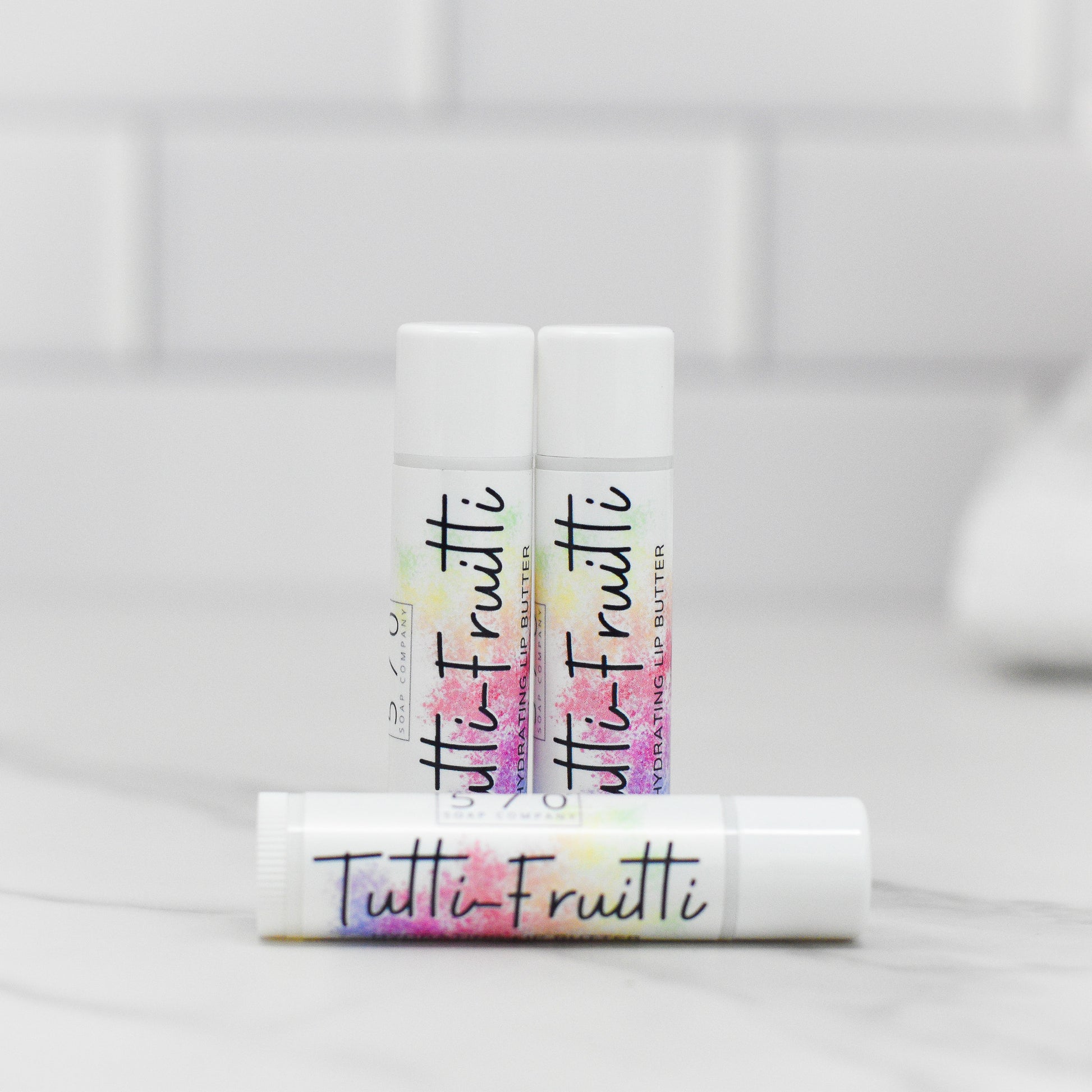 Three tubes of Tutti-Frutti Hydrating Lip Balm by 570 Soap Co. are showcased, with one tube lying flat and two standing upright. The vibrant labels stand out against a pristine white background with understated hints of a tiled wall. Made with natural ingredients, these lip balms provide luxurious care for vibrant and healthy-looking lips.