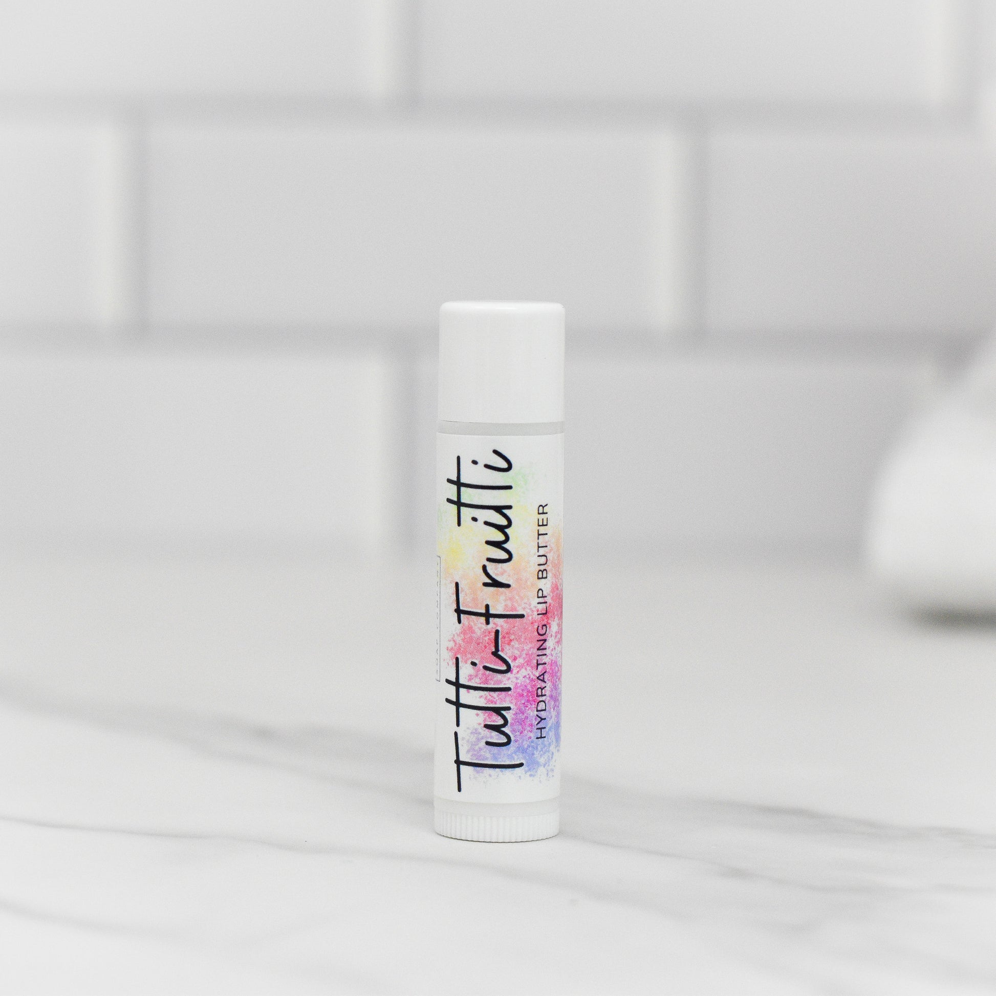 A tube of Tutti-Frutti Hydrating Lip Balm from 570 Soap Co. stands vertically on a marble countertop, delivering lip care with natural ingredients. The colorful text on the label set against a white background matches the blurred white tile backdrop, creating a clean, minimalistic vibe.