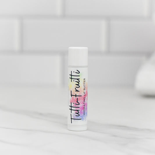 A tube of Tutti-Frutti Hydrating Lip Balm from 570 Soap Co. stands vertically on a marble countertop, delivering lip care with natural ingredients. The colorful text on the label set against a white background matches the blurred white tile backdrop, creating a clean, minimalistic vibe.