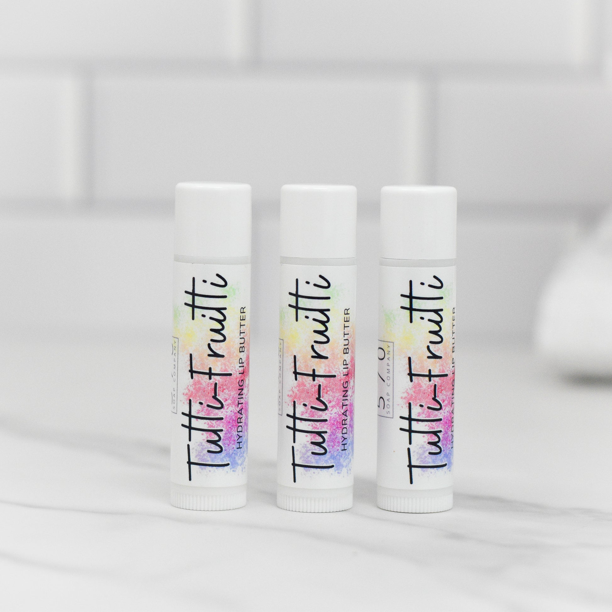Three white tubes of 570 Soap Co. Tutti-Frutti Hydrating Lip Balm, known for its natural ingredients, stand upright on a smooth, white surface. The background showcases a softly blurred white tiled wall, emphasizing the premium quality of this lip care product.