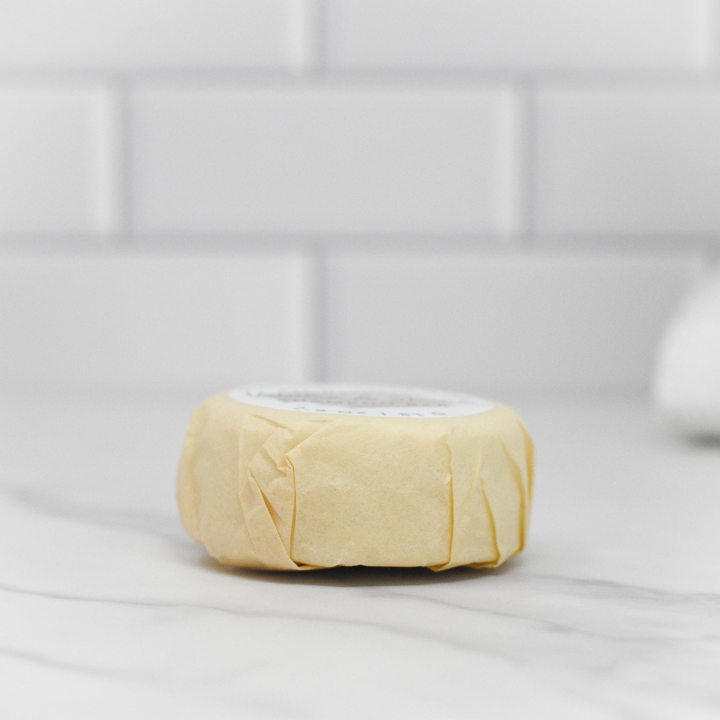 A small, round package of 570 Soap Co.'s Volume & Shine Hair Shampoo Bar - All Natural wrapped in beige paper sits on a white marble surface, with a white subway tile backsplash in the background.