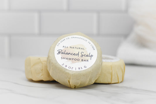 Three wrapped 570 Soap Co. Balanced Scalp Hair Shampoo bars, each 2.9 oz (81 g), are infused with tea tree oil and stacked against a white tile background. A towel is partially visible in the background.