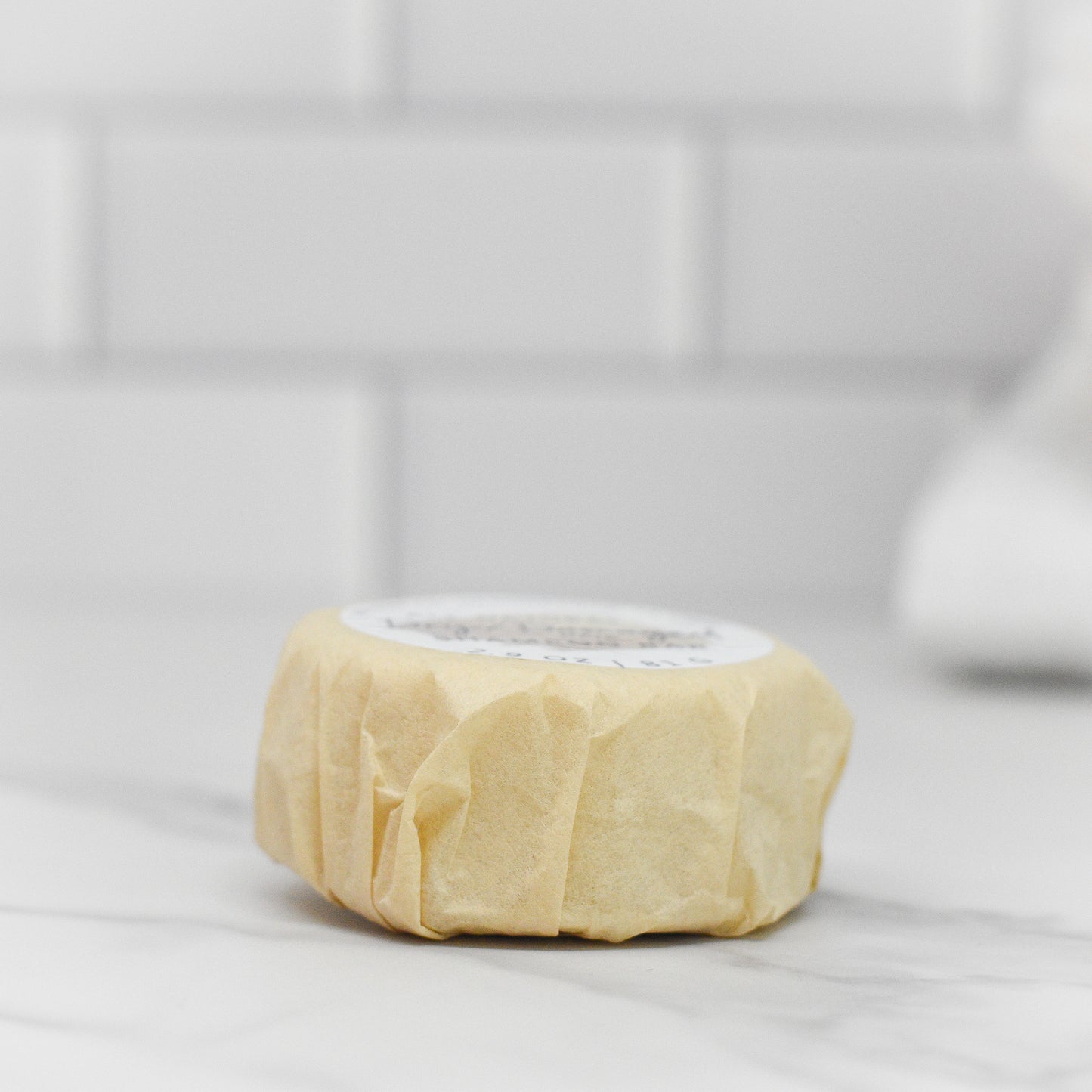 A round, beige shampoo bar from 570 Soap Co. rests on marble with a white tile backdrop. Wrapped in paper and infused with tea tree oil, the Balanced Scalp Hair Shampoo Bar is perfect for scalp balance.