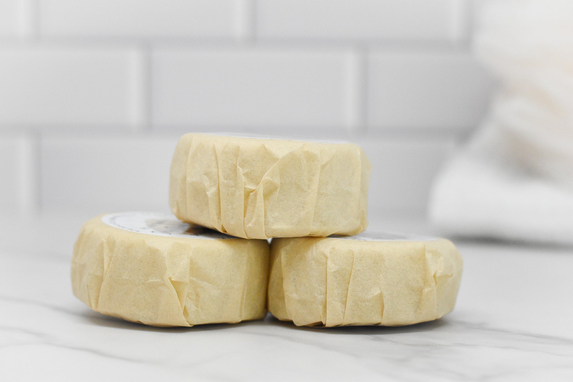 Three wrapped soap bars are stacked on a marble countertop with a white tiled backsplash in the background, accompanied by the 570 Soap Co.'s Curly Hair Shampoo Bar - All Natural to ensure frizz-free curly hair care.