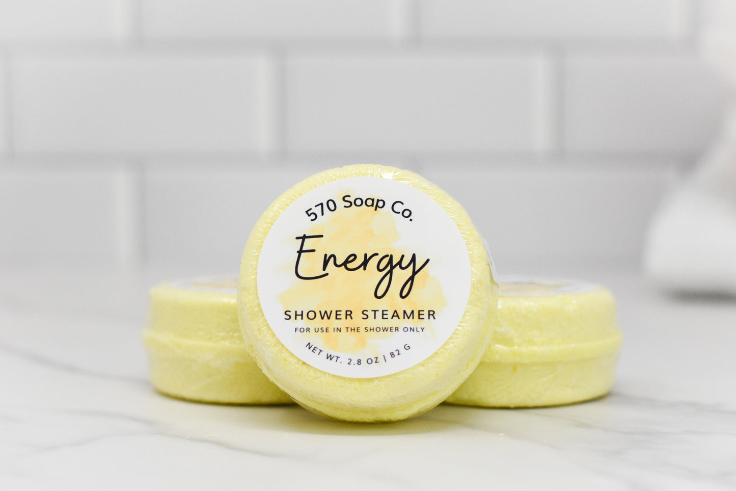 A stack of yellow Energy Shower Steamers from 570 Soap Co., infused with citrus essential oils and menthol, rests on a white surface. The front steamer's label indicates it is for shower use only and weighs 2.8 oz (82 g). In the background, pristine white tiles enhance the serene atmosphere.