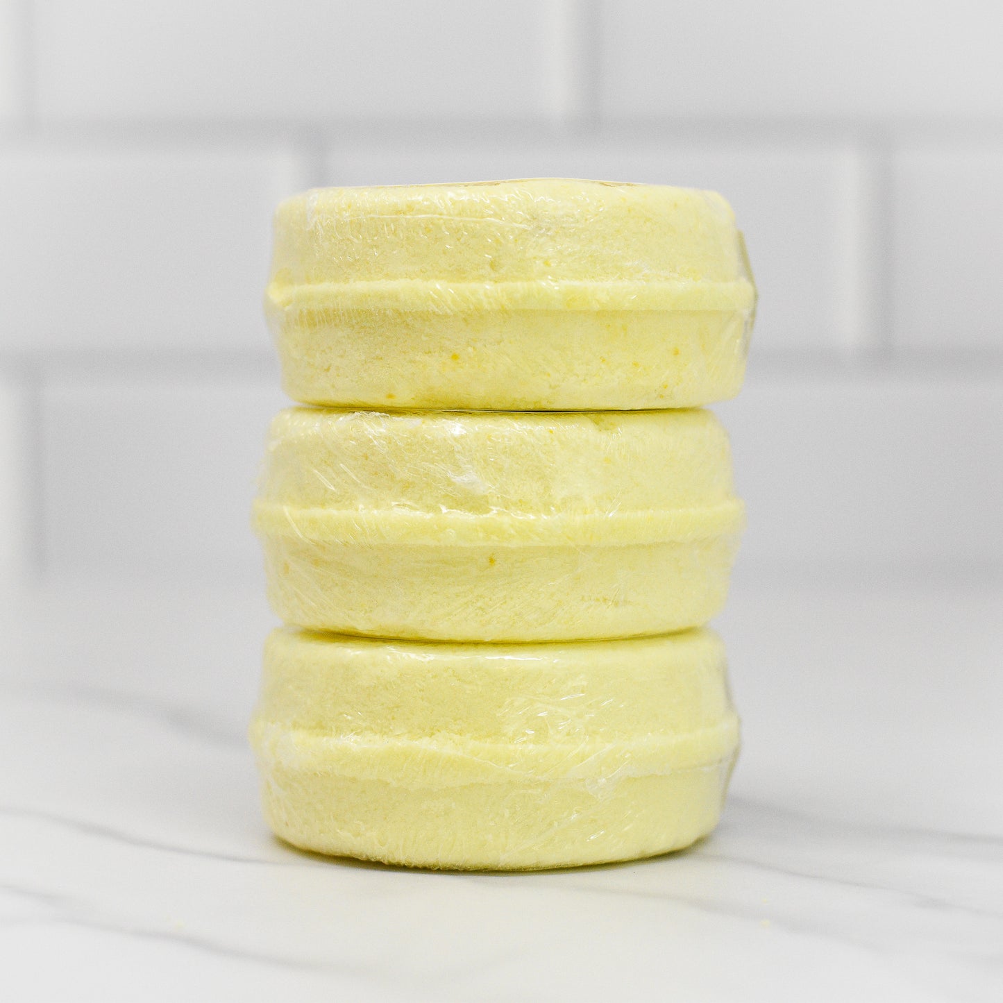 Three round soap bars from 570 Soap Co.'s Energy Shower Steamer - Citrus collection, infused with revitalizing citrus essential oils, sit on a marble surface wrapped in transparent plastic. The white tiled wall in the background highlights the vibrant and invigorating aura of these yellow soap bars.