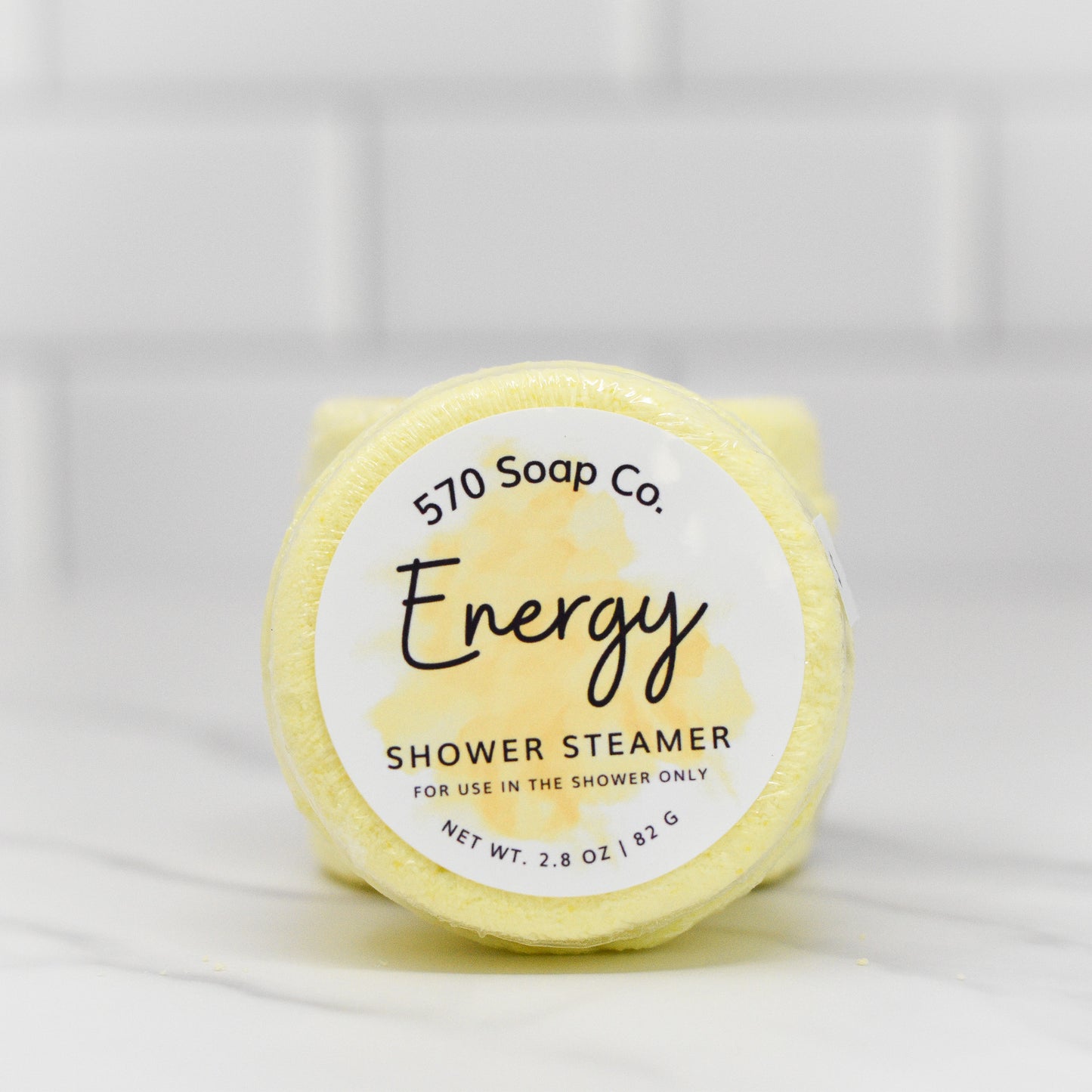 A round, yellow Energy Shower Steamer - Citrus from 570 Soap Co., infused with invigorating menthol and citrus essential oils, is set against a white tiled background. The packaging specifies shower use only and weighs 2.8 oz or 82 g, making it perfect for an energizing start to your day.