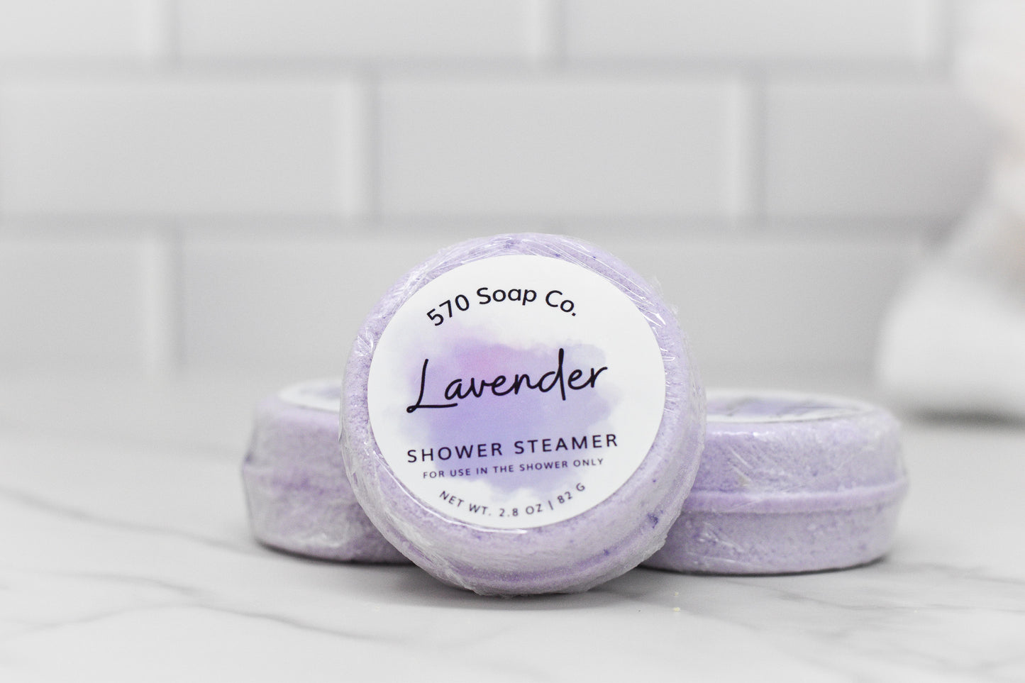 A set of Relax Shower Steamer - Lavender from 570 Soap Co. is artfully displayed against a white tiled background. The label on the front indicates that it offers a soothing shower experience. For shower use only. Net wt. 2.8 oz/1.16 g.